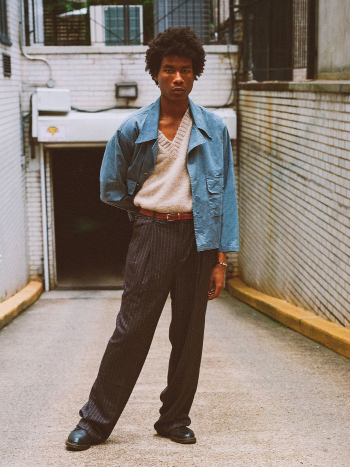In an alleyway, someone stands confidently in a Patina Work Jacket by Found over a beige sweater and pinstripe pants, embodying a timeless vintage workwear flair.