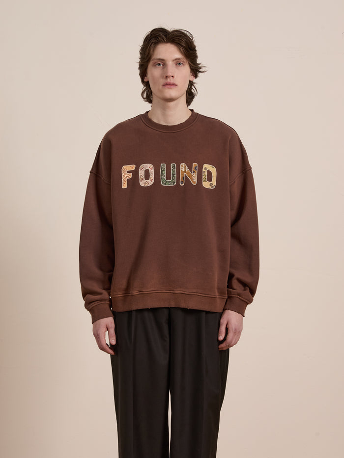 A person with medium-length hair wears the FOUND Patterned Logo Crewneck sweatshirt in brown against a neutral background.