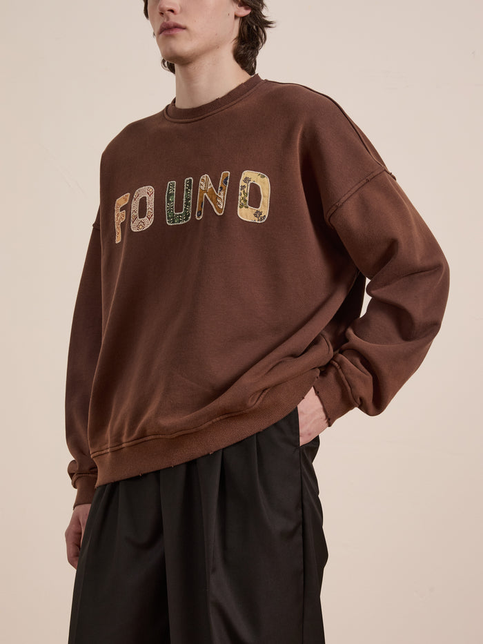 A person wearing a Patterned Logo Crewneck from FOUND, featuring brown patterns and appliqué "FOUND" lettering, paired with black pants.