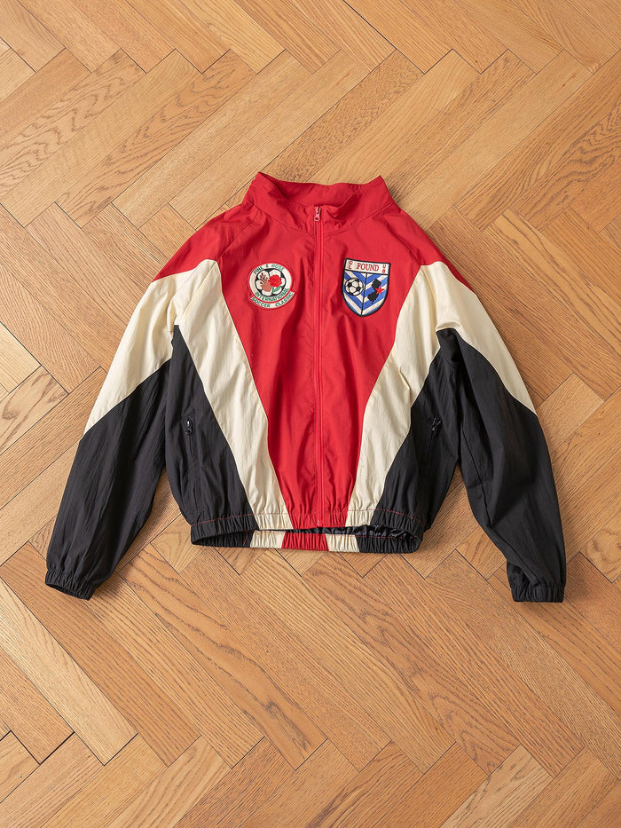 The Pine and Rose Track Jacket by FOUND, in red, beige, and black with two embroidered patches (a round text patch and a shield), offers unisex sportswear style and versatility. Displayed on a wooden floor.