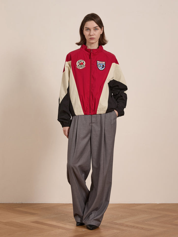 A person wearing the FOUND Pine and Rose Track Jacket with badges, paired with gray wide-leg trousers, stands on a wooden floor against a plain wall.