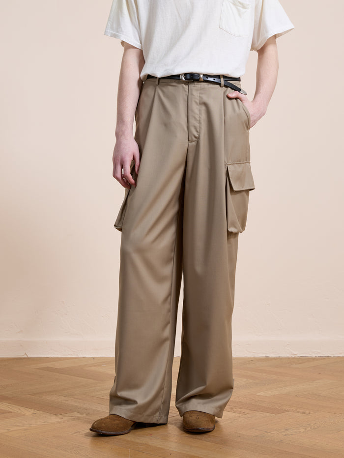 A person wears FOUND Pleated Cargo Pants—beige, relaxed fit with side pockets—paired with a white t-shirt, black belt, and brown shoes, standing on a wooden floor against a plain background.