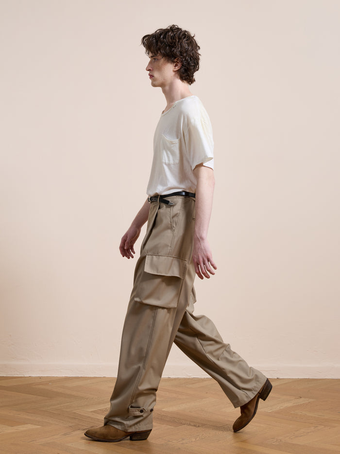 A person with curly hair walks indoors, wearing a white t-shirt, FOUND Pleated Cargo Pants in beige, a black belt, and brown shoes against a plain, neutral background.