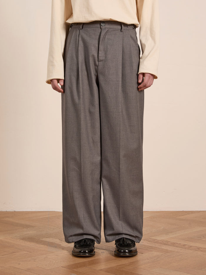 A person dressed in a beige long-sleeve shirt and FOUND's Pleated Trousers, featuring high-rise, double pleats, and adjustable waist tabs, is standing on an indoor wooden floor.
