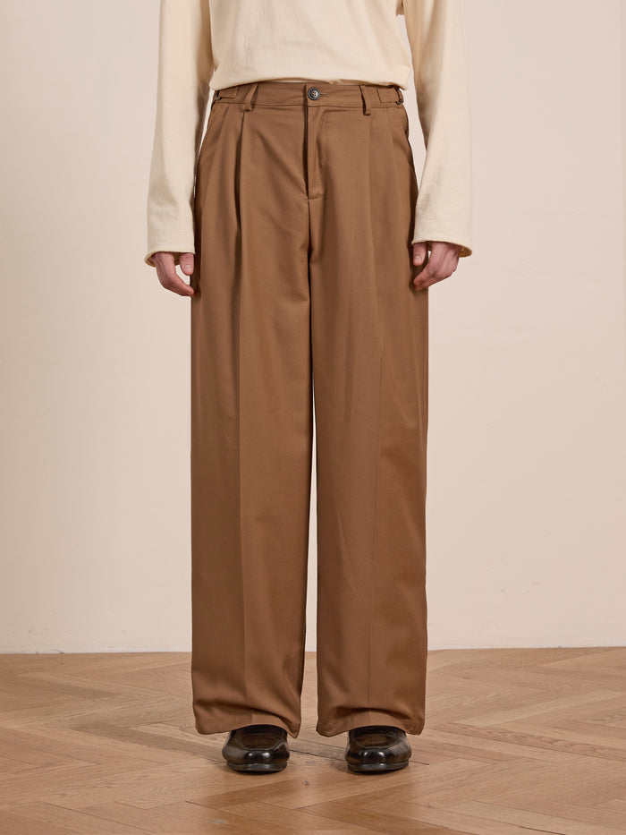 On a wooden floor, someone dons FOUND's pleated, relaxed-fit wide-legged brown trousers, paired seamlessly with a long-sleeved white top and sleek black shoes.
