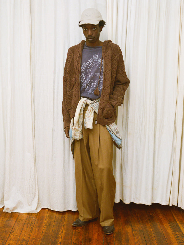 A person in FOUND Pleated Trousers, a light cap, brown knit cardigan, printed tee, and plaid shirt tied at the waist stands on a wooden floor before white curtains.