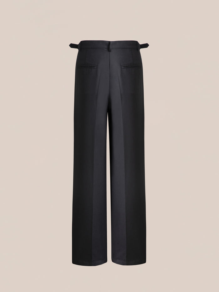 The Found Pleated Trousers, featuring a black wide-leg design with double pleats, adjustable waist tabs, and back pockets, are shown from the rear against a plain beige background.