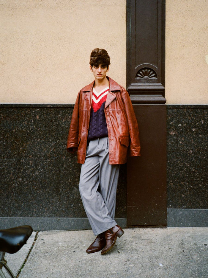 A person stands against a wall in a relaxed fit, wearing a brown leather jacket, patterned sweater, and high-rise gray Pleated Trousers by FOUND. Brown shoes complete the look, with a bicycle seat peeking into view.