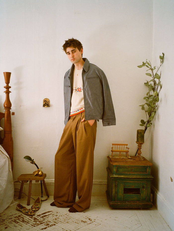 Amid a white wall, someone dons a gray jacket and FOUND's brown Pleated Trousers. A plant enhances the green cabinet as shoes sit near an elegant chair on the spotless floor.