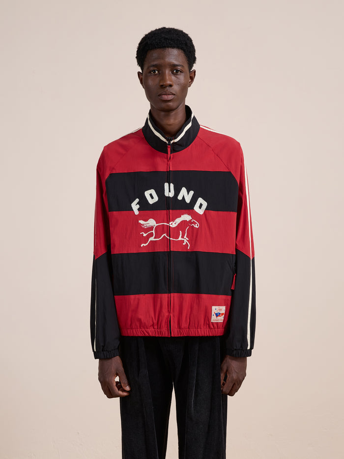 A person in a FOUND Racing Striped Windbreaker featuring "FOUND" and a running animal graphic on the front, styled like vintage sportswear, stands against a plain background.