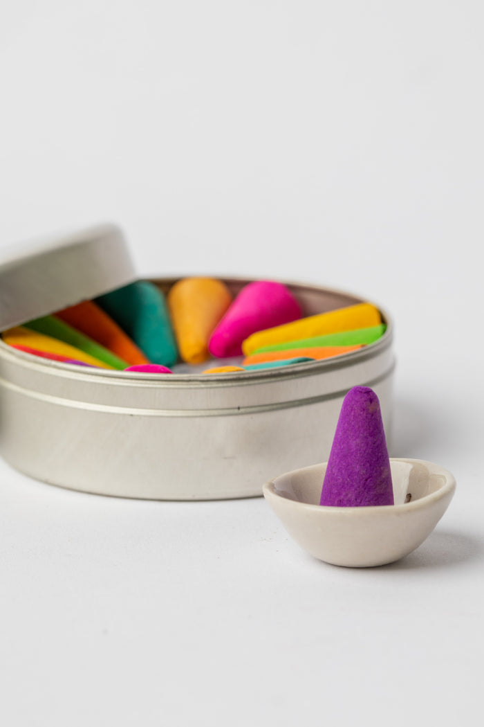 Purple incense cone displayed in ceramic holder beside open tin of rainbow incense cones