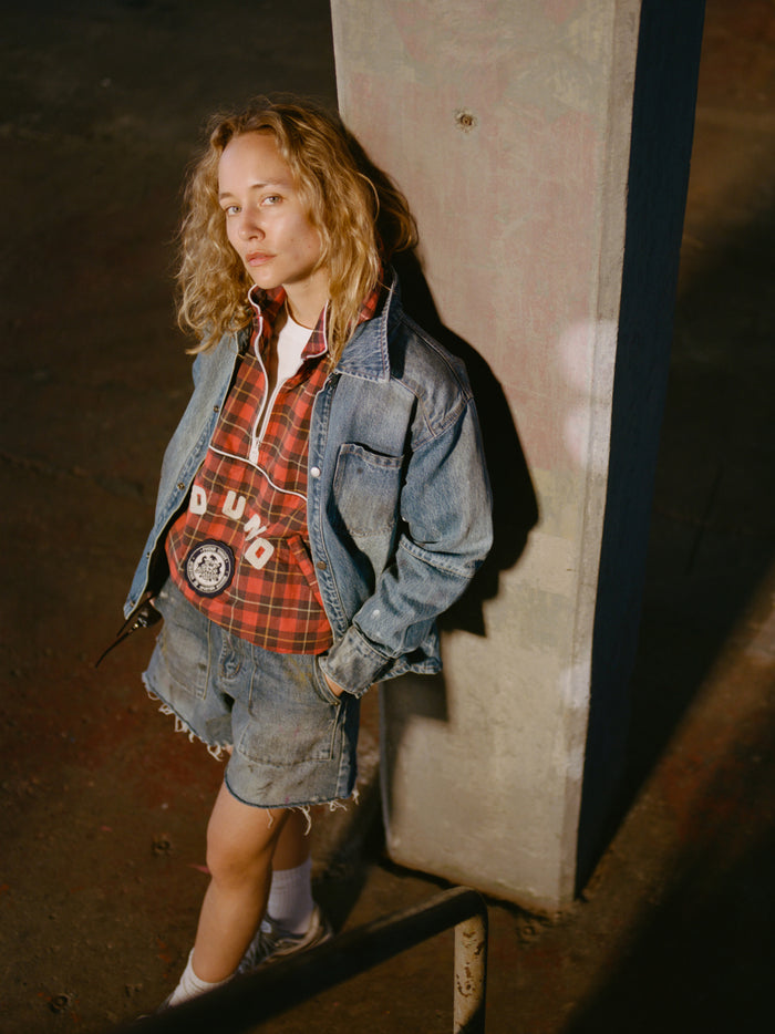 A person in a FOUND Red Plaid Track Jacket stands by a concrete pillar in a dimly lit indoor space, styled with denim shorts and white socks.