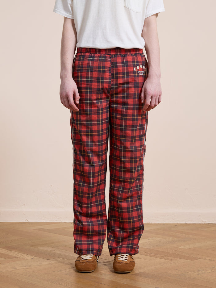 A person wearing Found Red Plaid Track Pants and a white short-sleeve t-shirt stands on a wooden floor against a plain beige wall.