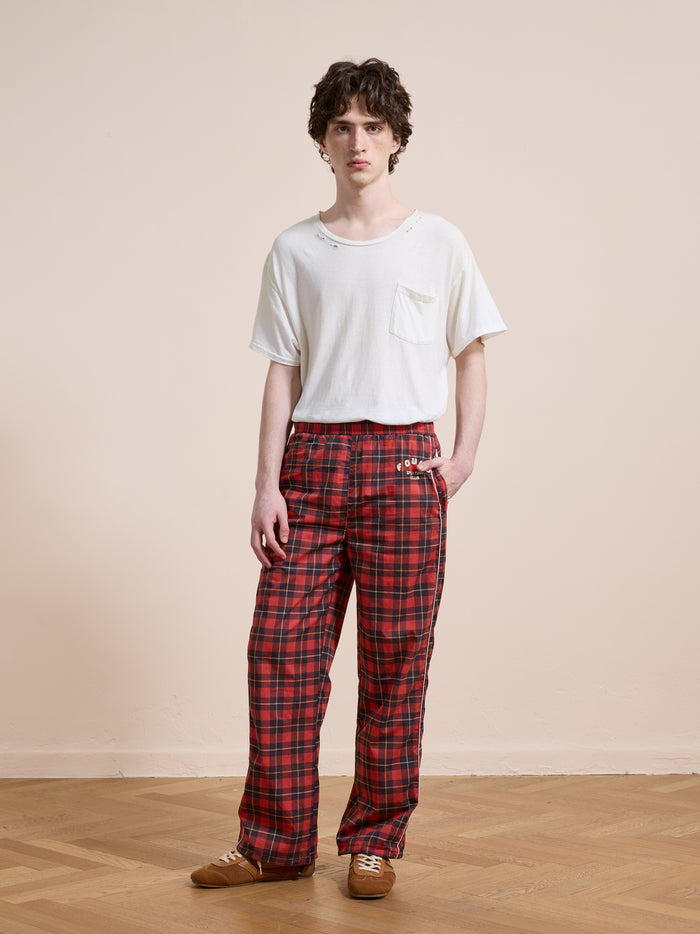 A person stands indoors against a beige wall and wooden floor, wearing Found's Red Plaid Track Pants with a relaxed fit, paired with a white pocket t-shirt and brown shoes.