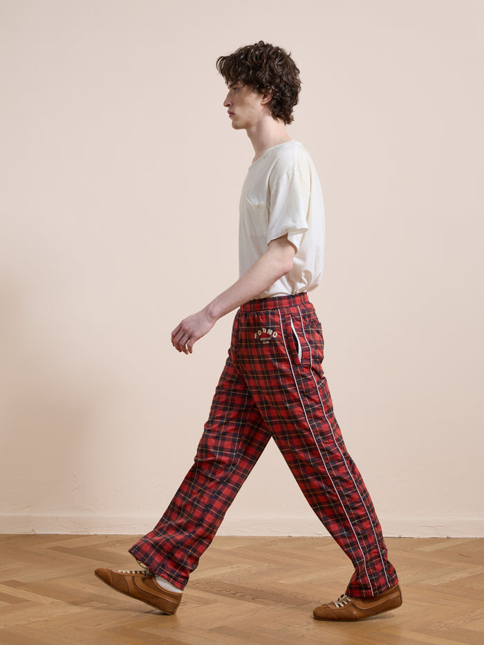 A person wears Found's Red Plaid Track Pants with a white T-shirt while walking on a wooden floor against a plain light-colored wall.