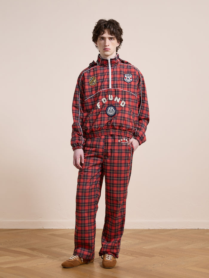 A person stands indoors wearing Found's Red Plaid Track Pants and a matching jacket with patches and text, paired with brown shoes, set against a plain beige background.