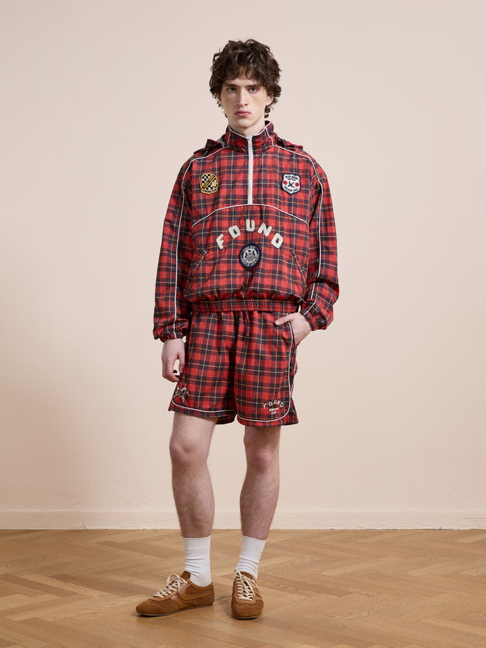 A person stands indoors against a beige wall and wooden floor, wearing Found Red Plaid Track Shorts and matching plaid jacket, styled with white socks and brown sneakers. The unisex relaxed fit creates a vintage athletic vibe.