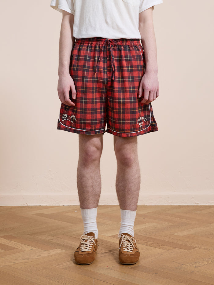 Indoors, a person wears a white t-shirt, Found Red Plaid Track Shorts (unisex relaxed fit with embroidery), white socks, and brown sneakers on a wooden floor.