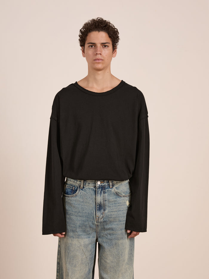 A person stands against a plain background, wearing a relaxed fit, unisex FOUND Reversed LS Tee in black paired with blue jeans.