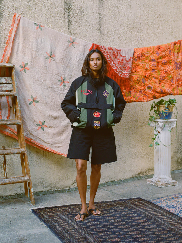 A person stands on a rug outdoors, wearing the FOUND Sporting Club Pullover Jacket in black and green with black shorts. Tapestries hang in the background, enhancing a vintage aesthetic alongside a wooden ladder and a plant on a pedestal.