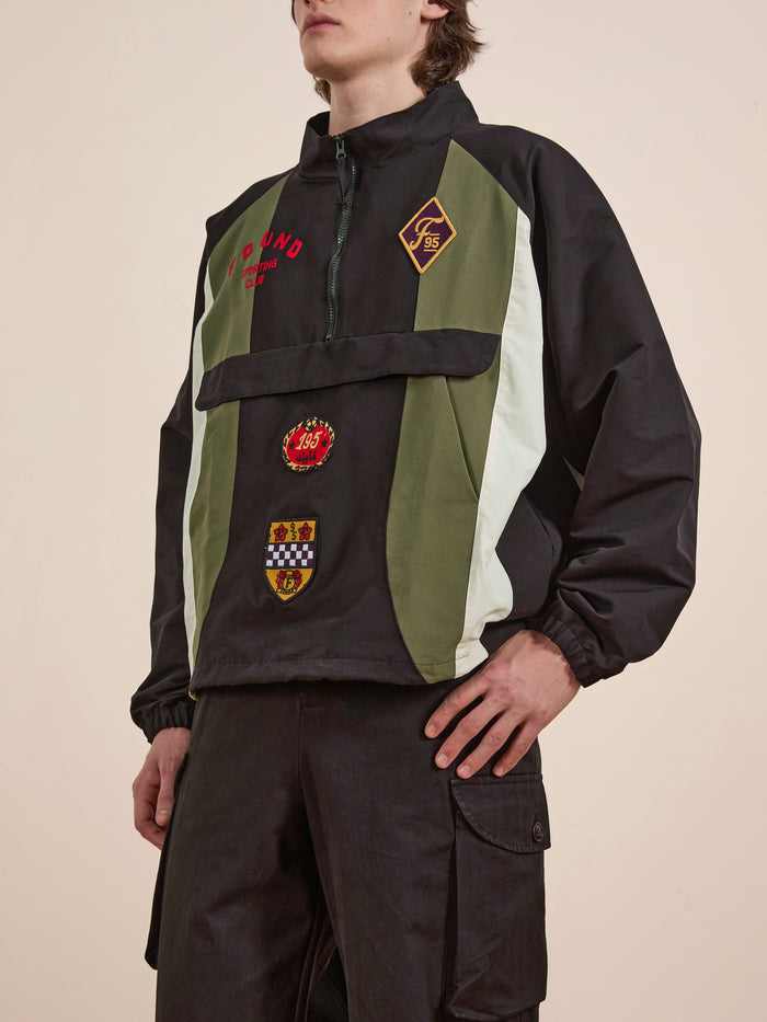 A person wearing FOUND's Sporting Club Pullover Jacket, featuring a vintage aesthetic in black, green, and white with red and multicolored patches, stands with one hand on their hip against a neutral background.