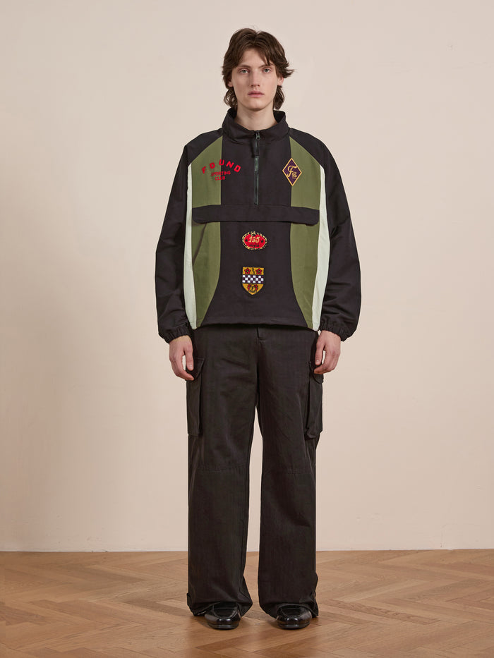 Indoors, a person exudes vintage flair in a FOUND Sporting Club Pullover Jacket in black and green with patches, alongside dark wide-leg pants and black shoes on polished wooden floors.