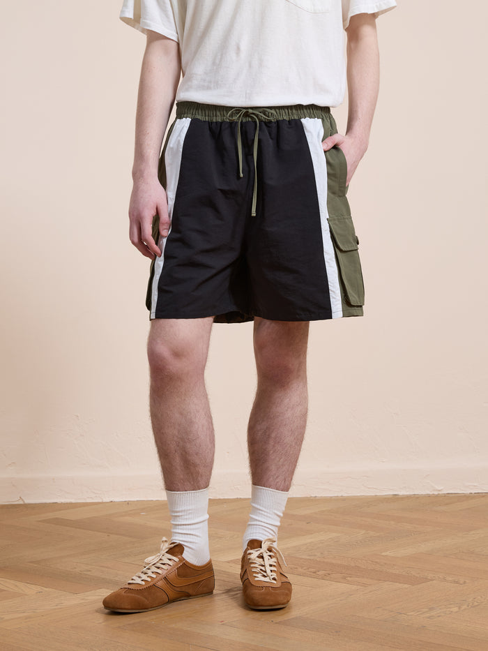 Someone wearing a white T-shirt and Found Sporting Club Track Shorts, featuring black, white, and green panels, along with white socks and brown sneakers, stands on a wooden floor.