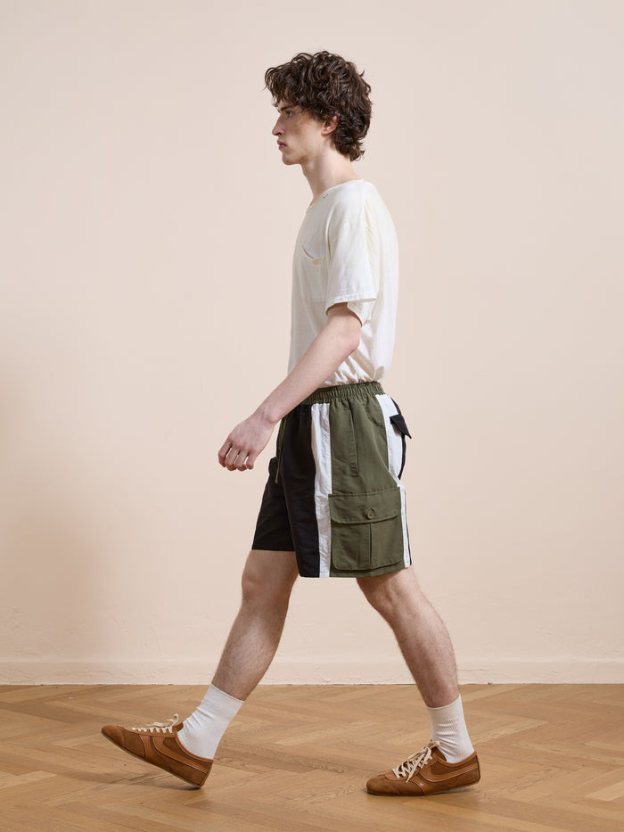 A man wearing a white t-shirt, Found Sporting Club Track Shorts in vintage green and white, white socks, and brown shoes walks indoors on a wooden floor with a plain beige wall in the background.