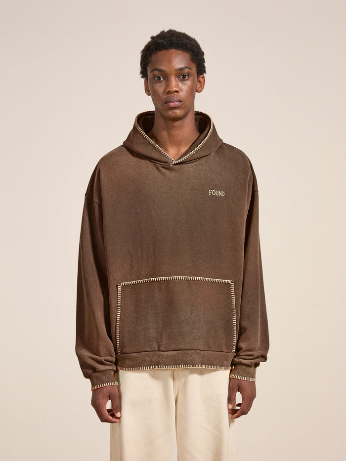 A person stands against a plain background wearing a vintage-style outfit, featuring a brown Stitched Border Distressed Logo Hoodie by FOUND, paired with light-colored pants.