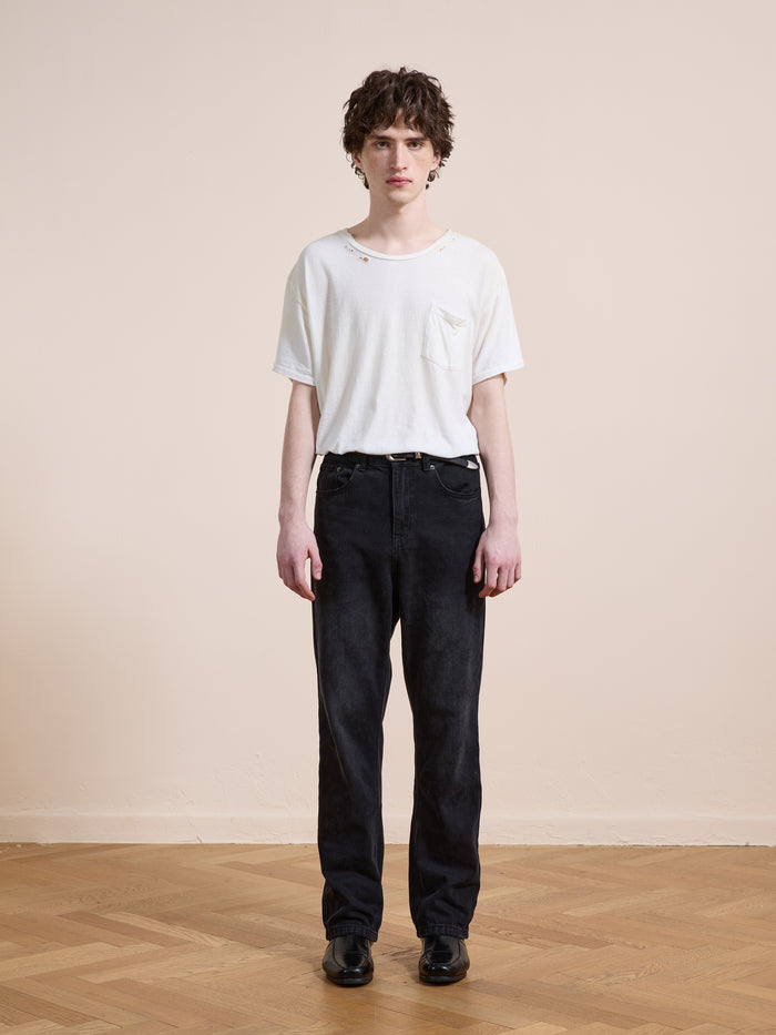 A person stands indoors on a wooden floor, wearing a white T-shirt with small holes, Found Straight Leg Jeans in black cotton denim, and black shoes, against a plain beige wall.