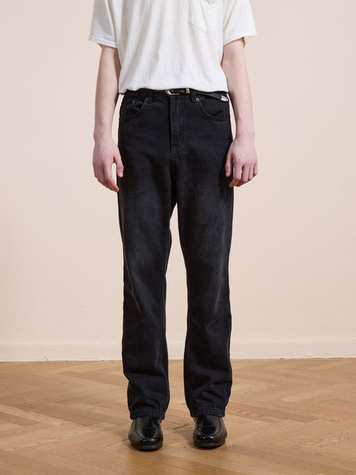 Someone wearing a white T-shirt tucked into Found Straight Leg Jeans in black cotton denim, styled with a black belt and shoes, stands on a wooden floor against a plain light-colored wall.