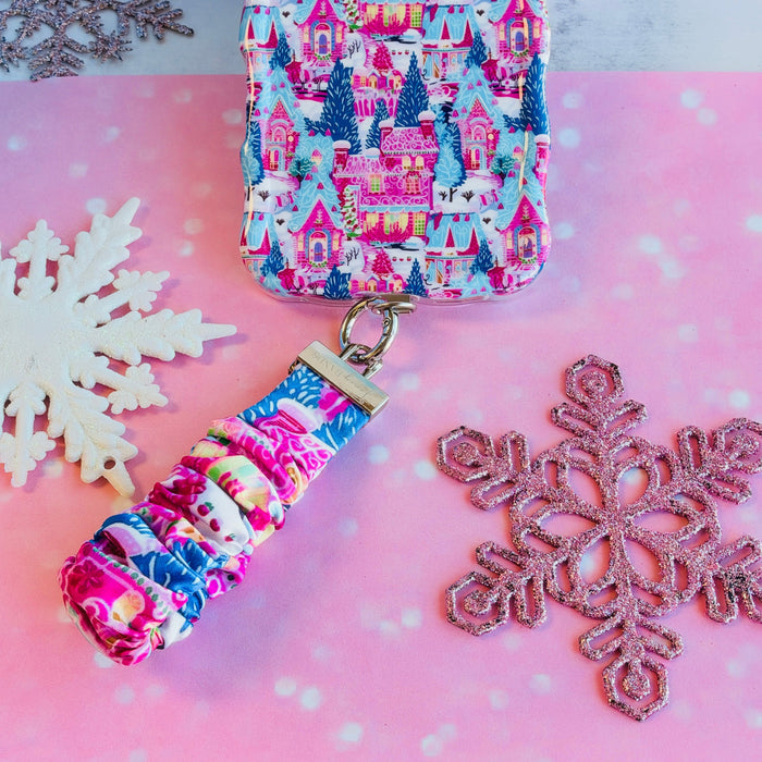 Sugarplum Square scrunchie phone wrist strap clipped below the matching Christmas village iPhone case on pink paper with glitter snowflakes