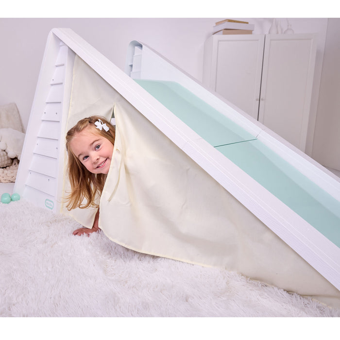 A young child with a white flower hair clip peeks out from inside a 'play tent' under a slide on a white shag rug.