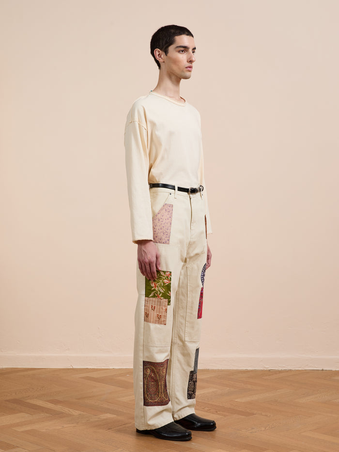 Indoors, a person wears a cream long-sleeve shirt and FOUND’s Tapestry Carpenter Canvas Pants, which feature vintage upholstery-inspired printed twill patchwork, styled with black shoes and a black belt.