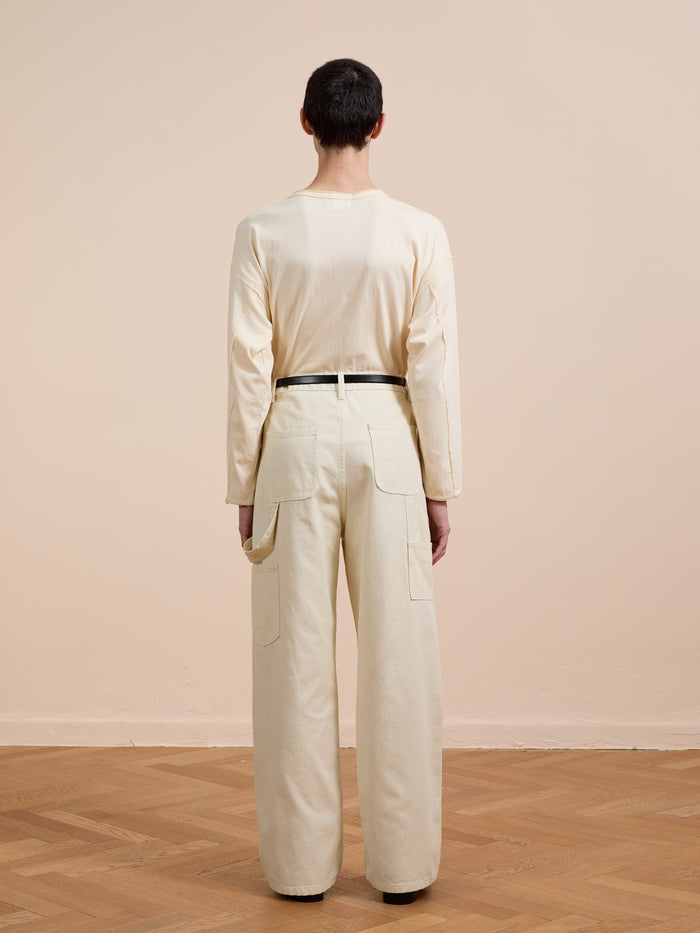 A person stands with their back to the camera wearing a cream long-sleeve shirt tucked into FOUND Tapestry Carpenter Canvas Pants, featuring a unisex relaxed fit, high waist, printed twill fabric, and large back pockets, on a wooden floor.