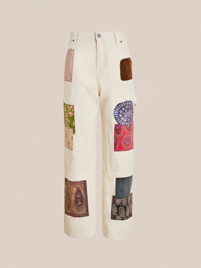 FOUND’s Tapestry Carpenter Canvas Pants are cream-colored unisex relaxed fit pants with multicolored fabric patches inspired by Pakistani quilt designs, sewn on the front and sides. Shown against a plain background.