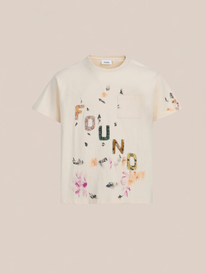 The FOUND Tapestry Logo Paint Tee is cream-colored, made from premium cotton, with a relaxed fit, chest pocket, and "FOUND" logo in mixed patterns plus floral and abstract prints.