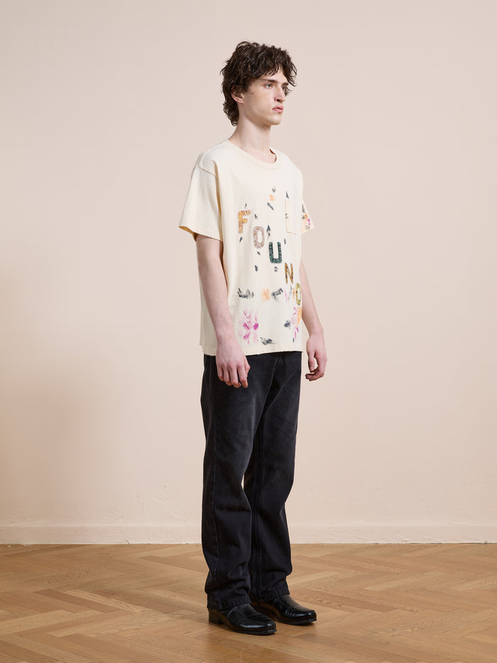 A person stands indoors on a wooden floor, facing slightly right against a beige wall, wearing FOUND's cream Tapestry Logo Paint Tee in premium cotton with colorful letters and black pants.