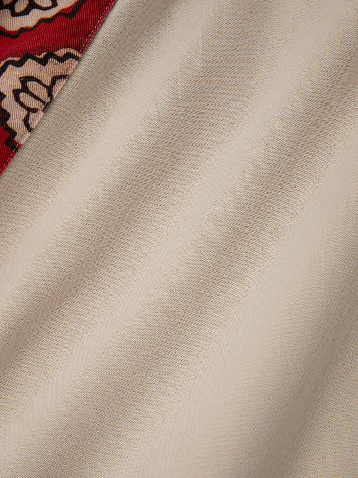 A detailed view of the Tapestry Patchwork Zip Up Hoodie by FOUND, showcasing its beige fabric intricately woven with South Asian tapestry patterns and highlighted by a red patterned border at the corner.