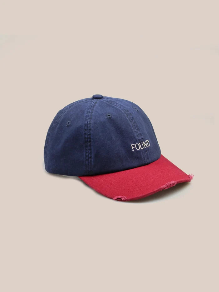 The Two-Tone Logo Cap by FOUND is a blue baseball cap with a navy cotton crown, a distressed red brim, "FOUND" embroidered on the front, and an adjustable velcro back strap for a perfect fit.