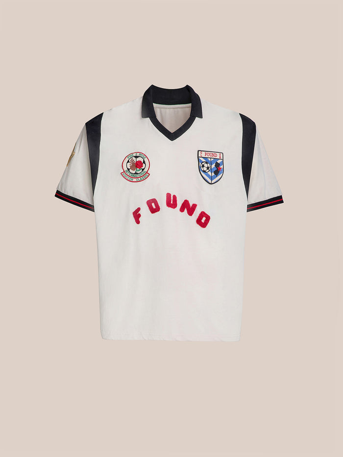 The Union Soccer Jersey by FOUND is a white jersey with a black collar and sleeves, featuring embroidered logos and "FOUND" in red across the front, offering a contemporary sportswear piece with vintage football vibes for those who love classic appeal.