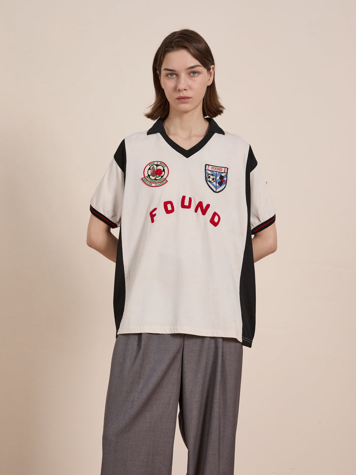 A person with short hair wears a Union Soccer Jersey by FOUND, a white shirt featuring patches and "FOUND" in red, evoking vintage football style. Paired with loose gray pants, they stand against a plain background exuding contemporary edge.