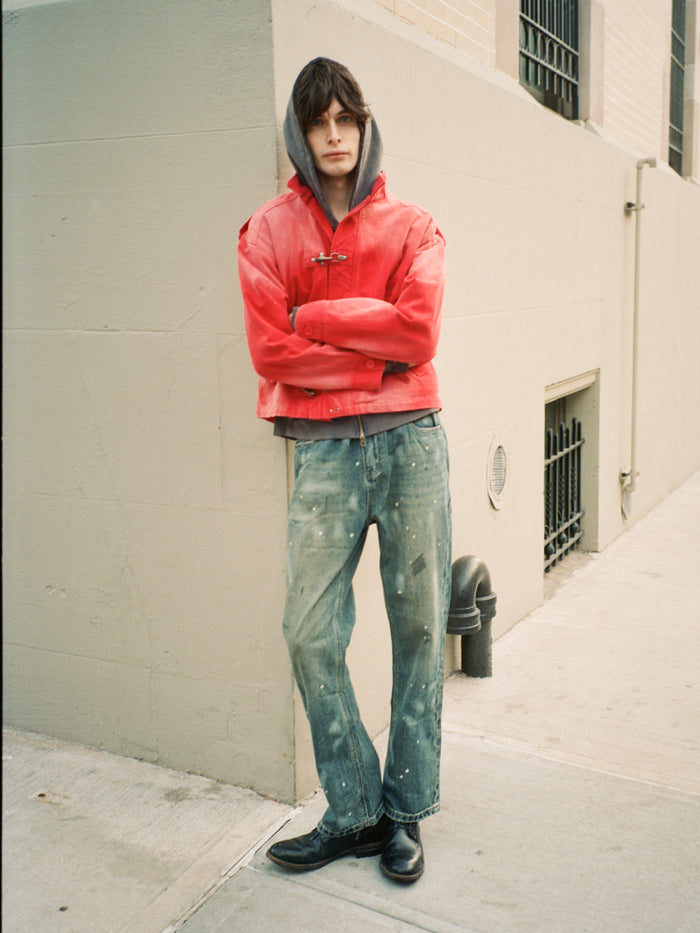A person stands on a sidewalk with arms crossed, wearing the FOUND Utility Clasp Work Jacket over a hoodie, paint-splattered jeans, and black shoes, leaning against a light-colored building wall.