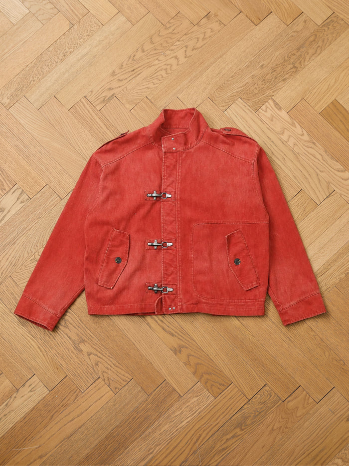 The FOUND Utility Clasp Work Jacket, a vintage-inspired red piece featuring long sleeves, front toggle closures reminiscent of fireman clasps, two flap pockets, and distinctive button details, is laid flat on a herringbone-patterned wooden floor.
