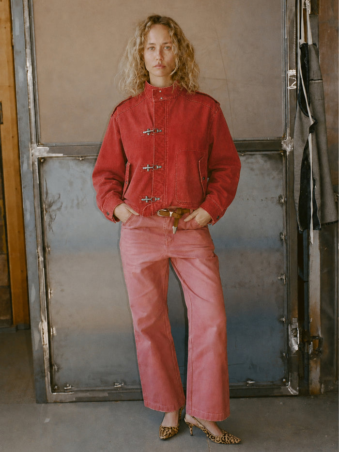 Person stands indoors wearing the FOUND Utility Clasp Work Jacket in vintage red with toggle closures, matching red pants, a brown belt, and leopard print heels, against a backdrop of metal and wood.