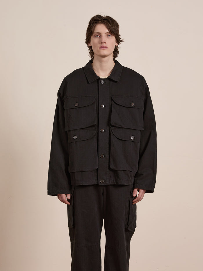 A person stands against a plain background, wearing a black Utility Herringbone Cargo Jacket by FOUND, featuring large pockets and a vintage look that seamlessly blends style and functionality.