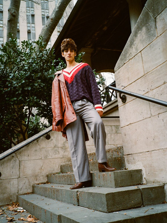 Wearing a FOUND V-Neck Cricket Sweater in navy and red, a person walks down stone steps with gray pants and brown shoes, holding a brown jacket. Trees and a building add elegance to the background.