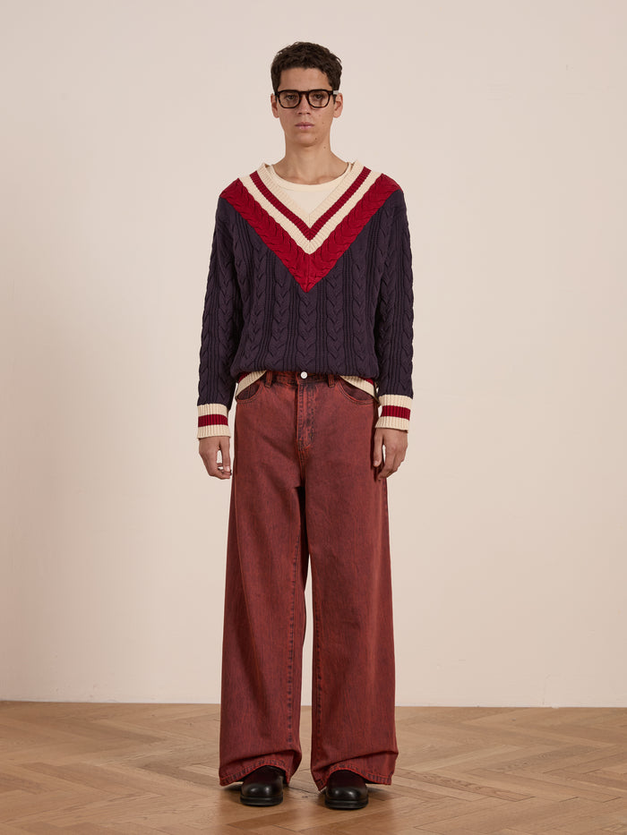 A person stands on a wooden floor wearing glasses and the FOUND V-Neck Cricket Sweater with red and white chevrons, paired with wide-legged red pants.