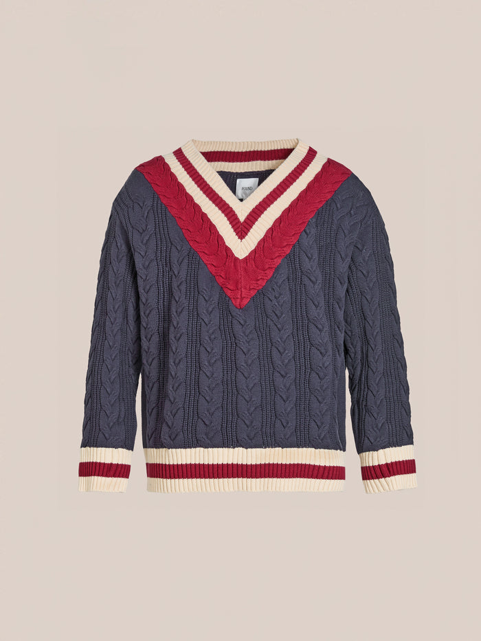 The FOUND V-Neck Cricket Sweater is navy with a cable-knit design, featuring a red and cream chevron pattern on the chest. It has matching sleeve trim and is crafted from soft cotton for ultimate comfort.