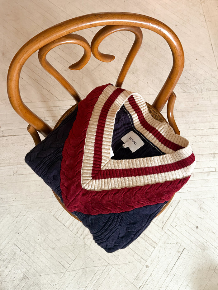 Two folded sweaters, a Found navy cable-knit and a red FOUND V-Neck Cricket Sweater featuring a white-striped collar, elegantly rest on a curved wooden chair.